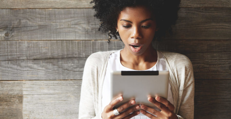 Portrait of beautiful shocked and astonished African woman with Afro hairstyle, holding tablet, reading surprising news on the Internet, opening mouth while posing against wooden wall background
