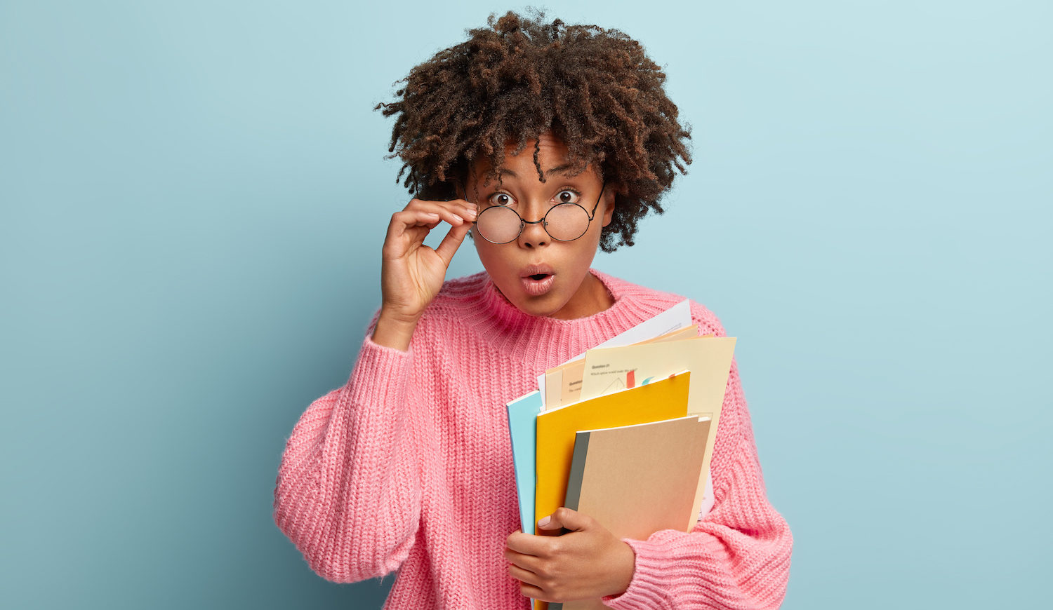 Image of surprised young teacher opens mouth as being speechless, holds some papers and notepad, touches frame of spectacles, looks shocked at camera, astonished by bad result of students exam