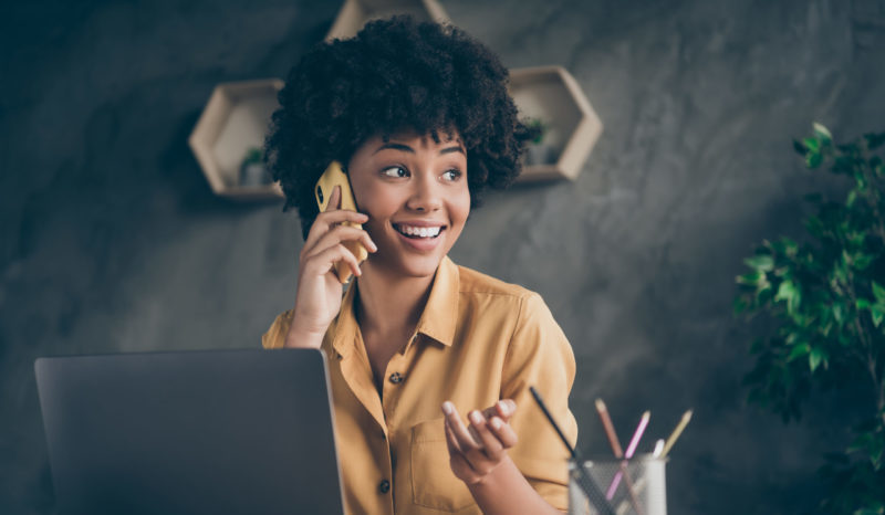 Photo of cheerful positive mixed-race pretty woman working as content manager arranging with her customers by phone smiling toothily